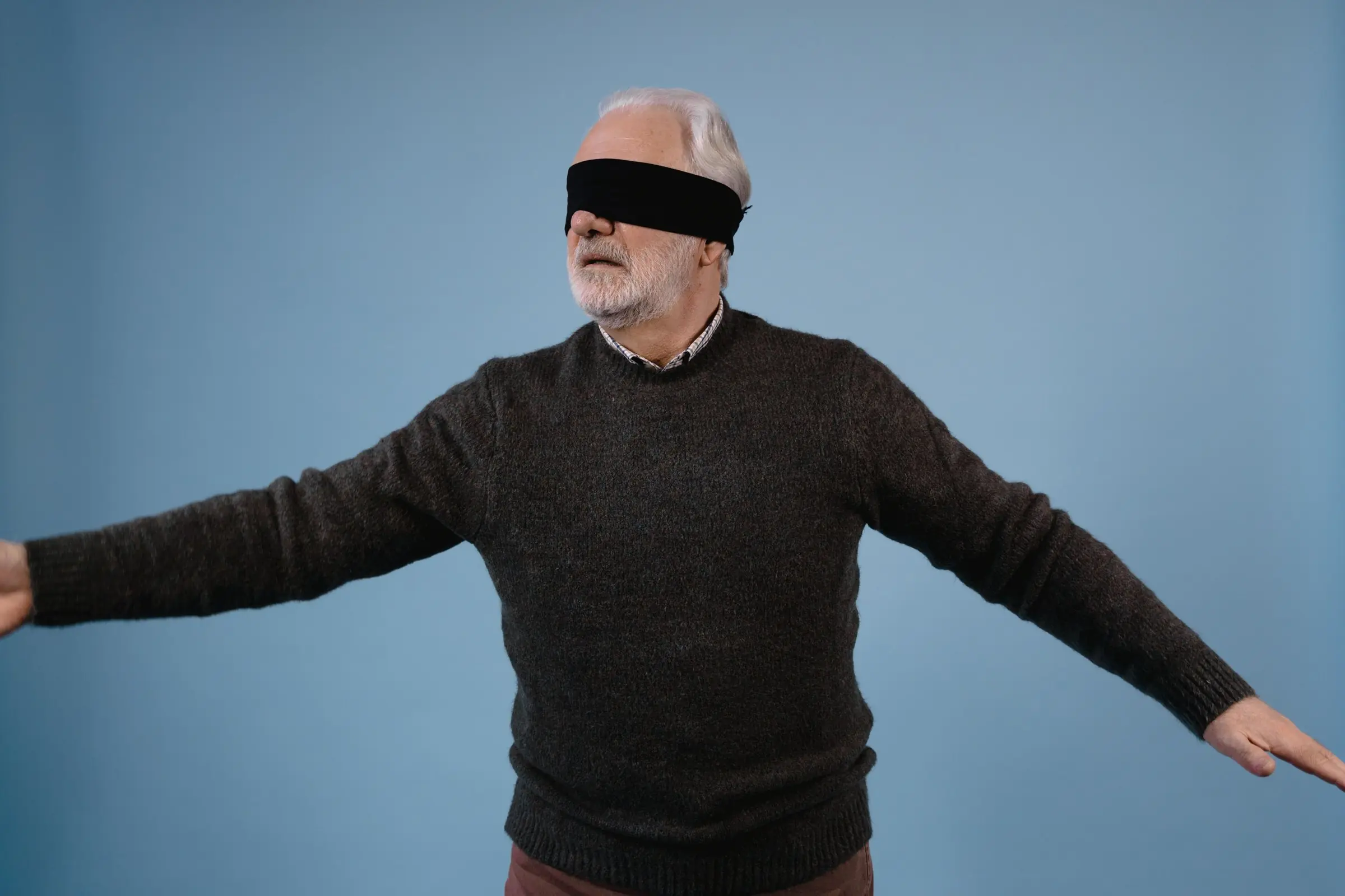 An older man wearing a blindfold, standing with his arms stretched out to keep his balance