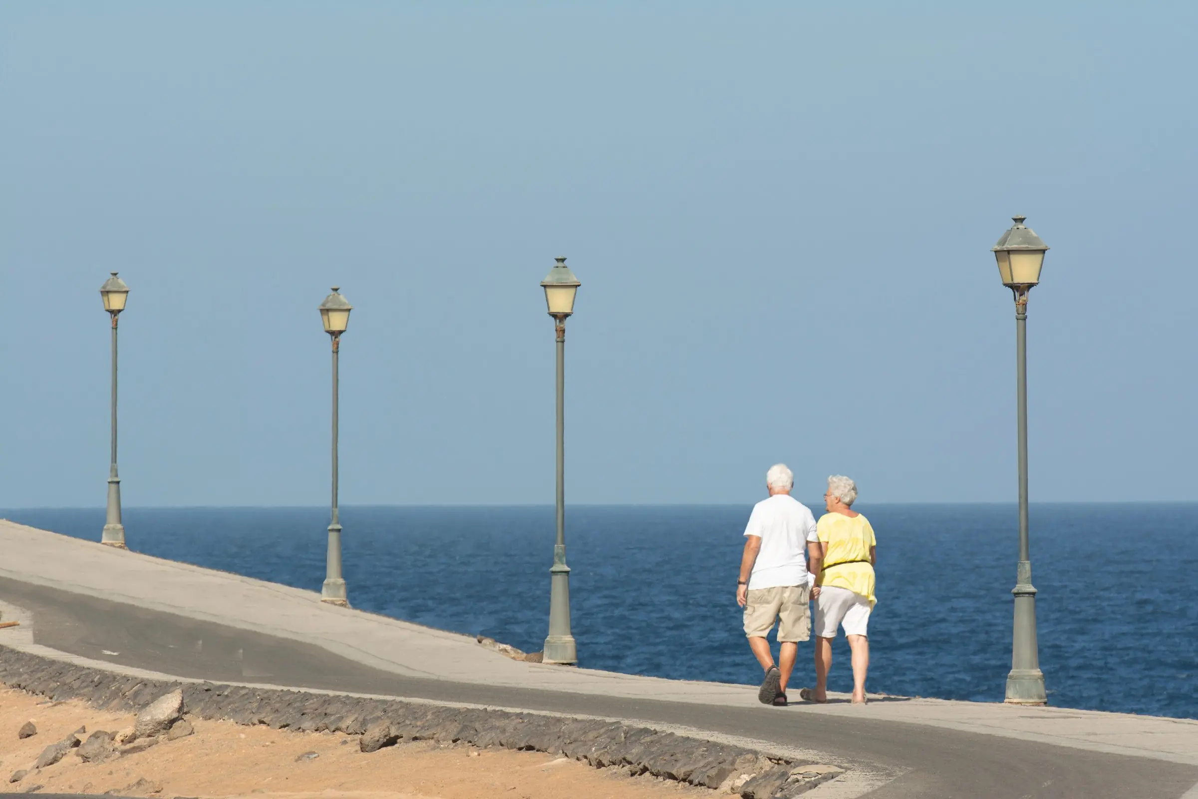 An older couple walking arm in arm along a seaside promenade beside a blue sea